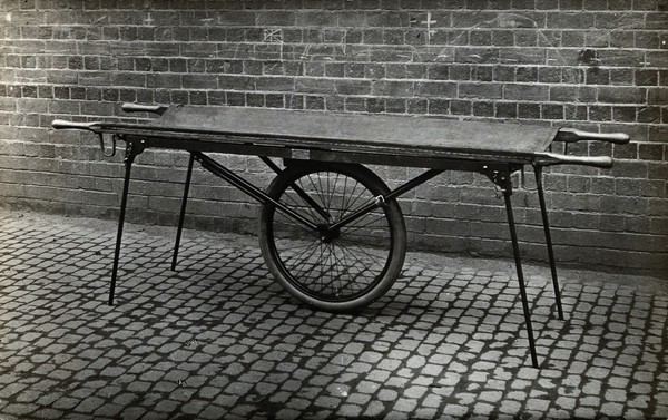 Closes' mono-wheel stretcher, in open position: side view, showing the stretcher resting on four fold-out legs, with one large central wheel in contact with the ground. Photograph, 1915/1918.