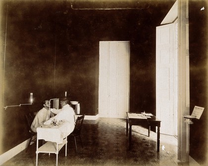 A tuberculosis patient undergoing an examination, Cuba: a doctor is shown examining the face of a patient, seated at a dispensary examination table. Photograph, 1902.