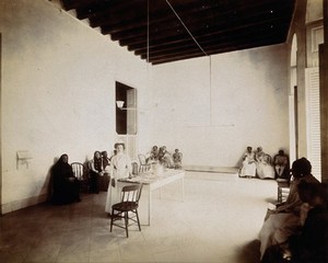 view Waiting room in a dispensary for the treatment of tuberculosis, Cuba: a nurse stands beside the reception table, surrounded by seated patients. Photograph, 1902.