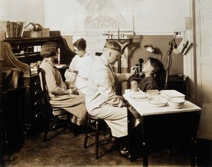 view A Pennsylvania State tuberculosis clinic: a male doctor is shown examining the tongue of a boy, while a female medical practitioner takes the pulse of a woman. Photograph, 1925/1935?.