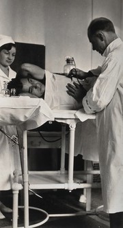Pneumothorax treatment, Cresson State Sanatorium, Pennsylvania: a doctor is shown adminstering the treatment to a male patient, who is lying on his side, watched over by a nurse. Photograph, 1920/1940?.