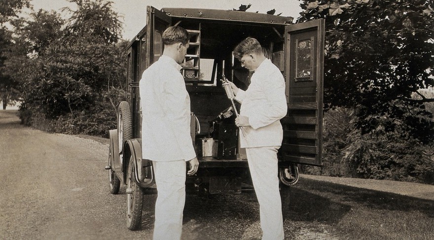 A mobile milk control field laboratory, Pennsylvania: two men wearing white suits take a large thermometer from the back of the van. Photograph, 1920/1930?.