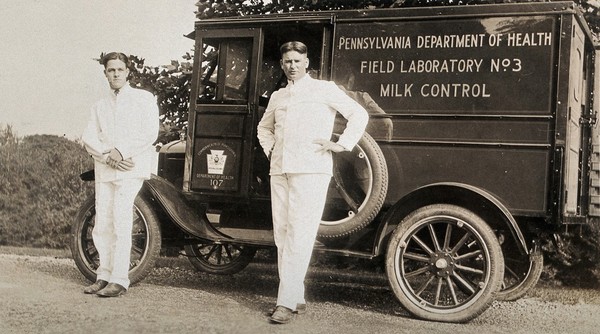 A mobile milk control field laboratory, Pennsylvania: two men wearing white suits stand beside their van. Photograph, 1920/1930?.