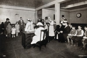 view Interior of a state clinic, Pennsylvania: a nurse is shown seated at a desk, while doctor measures a girl's height and other patients wait to be seen. Photograph, 1910/1930?.