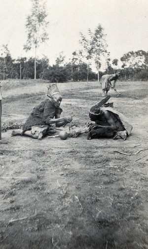 view Ankole, Uganda: two medicine men kneeling over the corpse of a small animal. Photograph by John Roscoe, 1919/1920.