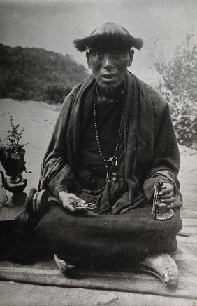 A man (a Buddhist monk?) sitting cross-legged wearing robes and beads and holding a bell. Photograph, 1910/1930.