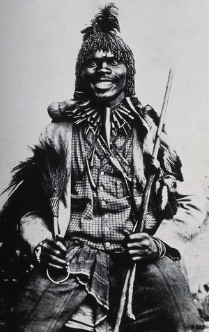 view Africa: a medicine man in ceremonial dress holding a staff and a whisk. Reproduction of a photograph, 1880/1900 (?).