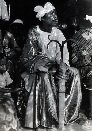 view Africa: a medicine man, seated, holding a wooden staff with metal prongs. Photograph (by Kurt Lubinski?), 1940/1960.