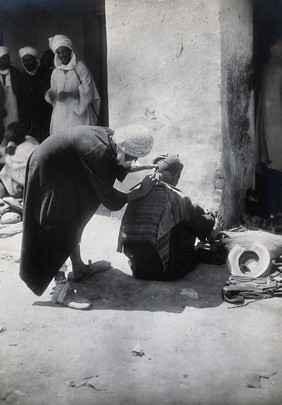 Sudan: a barber at work on a seated customer in the street. Photograph, ca. 1920.