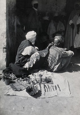 Sudan: a barber at work on a seated customer in the street, his tools spread out on the ground next to him. Photograph, ca. 1920.