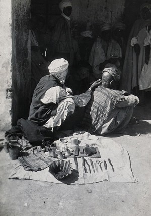 view Sudan: a barber at work on a seated customer in the street, his tools spread out on the ground next to him. Photograph, ca. 1920.