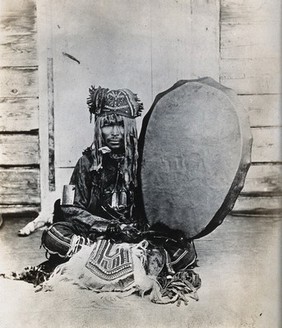 Siberia: a Tungus medicine man in ceremonial dress, seated holding a drum. Photograph, ca. 1920 (?) of a photograph by Marya Czaplicka, 1914/1915.