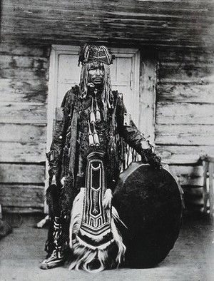 view Siberia: a Tungus medicine man in ceremonial dress, standing holding a drum. Photograph, ca. 1920 (?) of a photograph by Marya Czaplicka, 1914/1915.