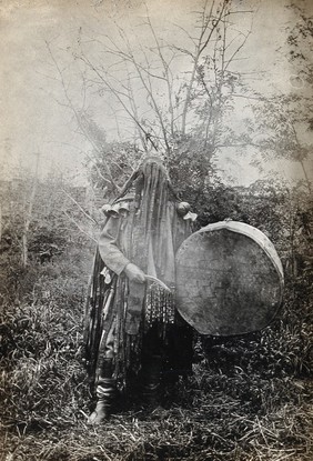 Minusinsk, Siberia: a Tartar medicine man or shaman in ceremonial dress with a covered face, holding a drum. Photograph, ca. 1920 (?) of a photograph, 1900/1915.