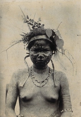 A young woman with extensive scarification on her face, wearing a hat of foliage: front view. Photograph.