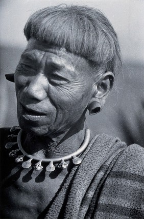 A man of the Konyak Naga tribe, with plugs distorting his ears and a necklace indicating the number of heads he has cut off. Photograph.