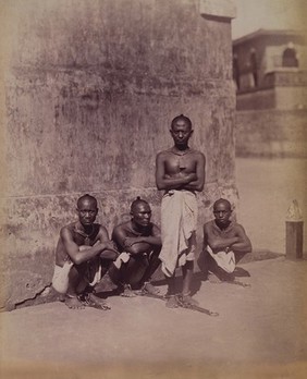 India: four convicted robbers with leg-irons on their feet and labels suspended around their necks. Photograph, 18--.