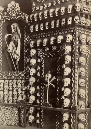 view Skulls and skeletons of friars arranged columns around the walls of a chapel. Photograph.