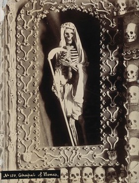 The skeleton of a friar in the role of Death as the reaper, surrounded by skulls, ribs and vertebrae, in a chapel. Photograph.