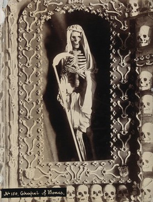 view The skeleton of a friar in the role of Death as the reaper, surrounded by skulls, ribs and vertebrae, in a chapel. Photograph.