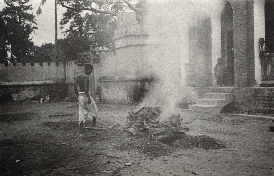 Calcutta, West Bengal: the dead body of a man burning on a funeral pyre; a young man standing by. Photograph.