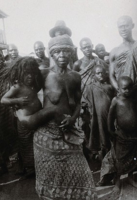 Kumasi, Ghana: a mother holding a child with unshaved hair; a group stands in the background. Photograph, 1920/1940 (?).