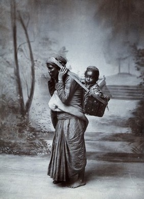 A woman a carrying a child in a basket on her back, supported by a strap around her head: studio portrait. Photograph, ca. 1900.