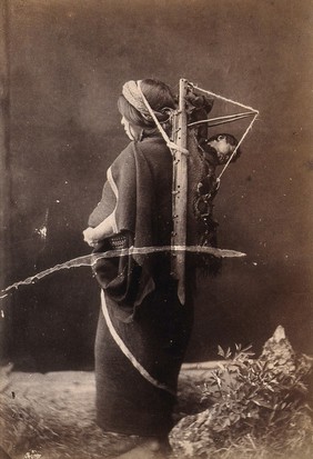 An Araucanian woman in Chile carrying a sleeping child on her back in a wooden carrier, supported by a strap around her head. Photograph, ca. 1913.