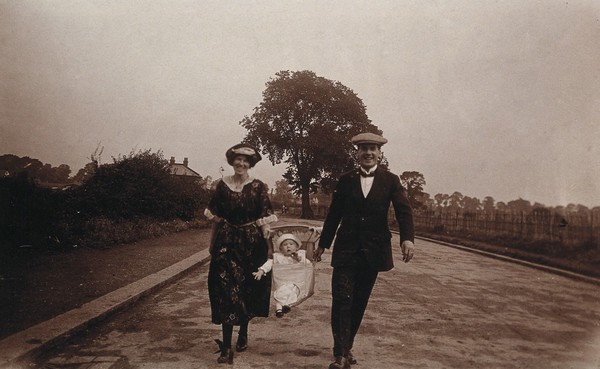 A baby in a carrier made from canvas sailor's trousers, being carried along the street by its parents in Southend-on-Sea, England. Photograph by Cyril Norwood, 1922.