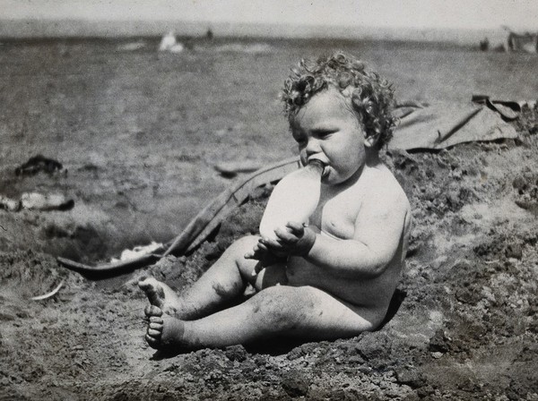 A baby drinking milk from a feeding bottle, seated naked on sand. Photograph, 1922.