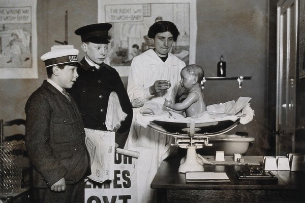 Ideal Home Exhibition, England, 1920: a rubber baby being used to demonstrate feeding methods, watched by two boys. Photograph, 1920.