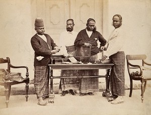 view Galle, Sri Lanka: jewellers in traditional dress displaying their jewellery on a table. Photograph, ca. 1900.