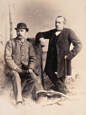 view Two American men: studio portrait: one seated on a tree stump, one standing, leaning on the tree stump. Photograph, ca. 1880.