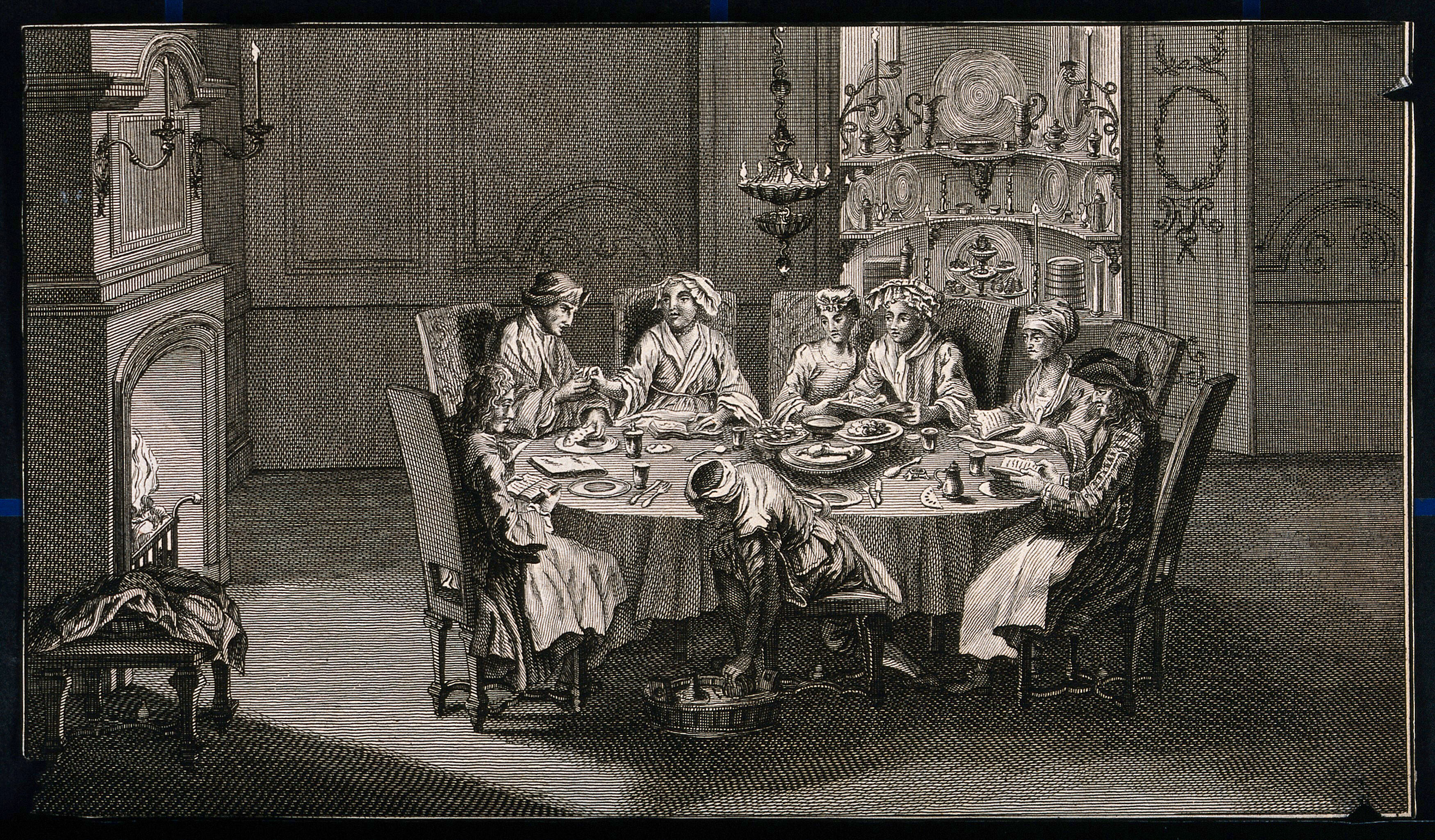A Group Of People Sit Around A Table Sharing A Meal And A Person In A Turban Sitting On A Stool Bends Down To Pick Something Out From A Tray On The