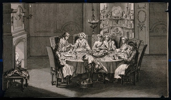 A group of people sit around a table sharing a meal and a person in a turban sitting on a stool bends down to pick something out from a tray on the floor. Engraving.