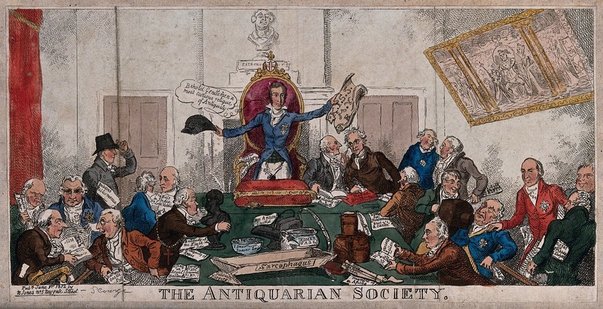 Antiquaries meeting: a man standing at the head of a table holds up a hat in one hand and a sheaf of papers in the other for the rest of the company to see; the table has busts and pots on it and the men are inspecting various items. Coloured etching.