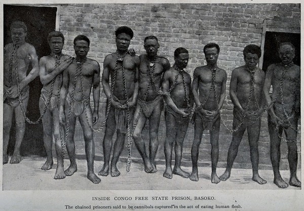 Nine male prisoners in the Congo standing against a wall joined by chains around their necks. Process print after W.E. Geil.