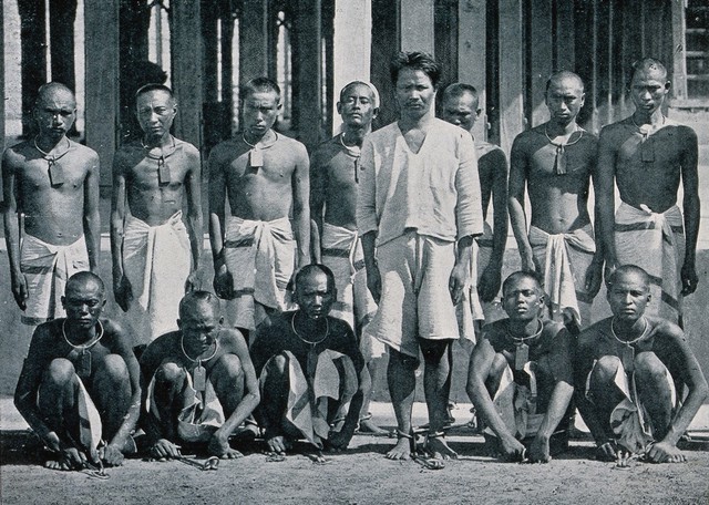 Thirteen male prisoners in India or Burma ("dacoits", bandits) wearing chains around their necks and ankles. Process print.