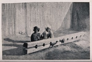 view Two Indian men seated on the ground, their left feet trapped in a pillory. Halftone.