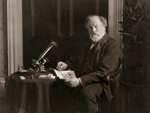 view A scientist, posed with his microscope, in a drawing room setting. Photograph by Elliott and Fry.