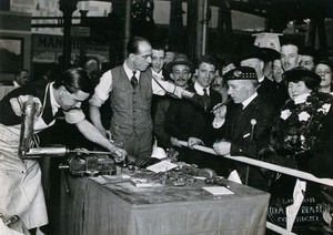 view Sir Harry Lauder, the Scottish entertainer, at a prosthesis stand at a Daily Mail exhibition, Olympia, 1921: one man with an arm prosthesis lights his pipe, a second demonstrates the equipment. Photograph, 1921.