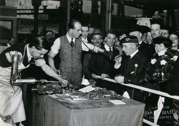 Sir Harry Lauder, the Scottish entertainer, at a prosthesis stand at a Daily Mail exhibition, Olympia, 1921: one man with an arm prosthesis lights his pipe, a second demonstrates the equipment. Photograph, 1921.