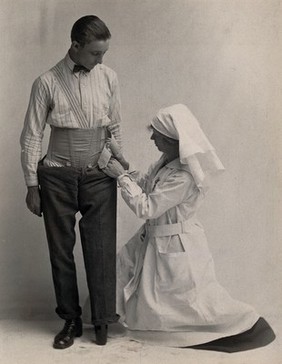 A man wearing a leg prosthesis aided by a nurse who kneels to adjust the straps. Photograph, 1914/1918.