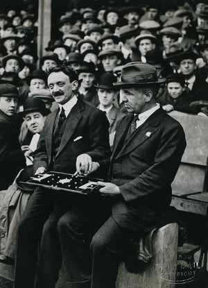 view A blind man at a football match in London, following the game using a board representing a pitch and players, assisted by the board's inventor. Photograph, ca. 1921.