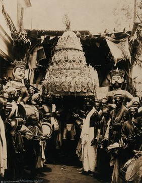 India: the procession of the Sacred Water Pot to avert cholera: the decorative over-sized pot is held aloft between two crowned models; musicians stand to each side. Photograph by Underwood and Underwood, ca. 1900 (?).