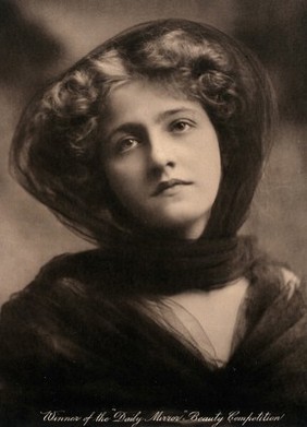 A young woman, winner of a beauty contest, wearing a large black hat with a trailing scarf round her throat.