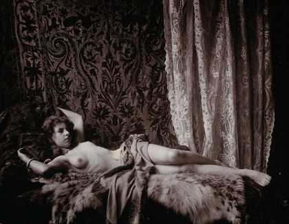 A young woman, posing semi-naked in a photographic studio, lying full-length on a fur-covered sofa or day-bed.