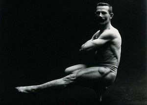 view A male bodybuilder wearing bathing trunks, striking a "Cossack dance" pose, in a studio setting.