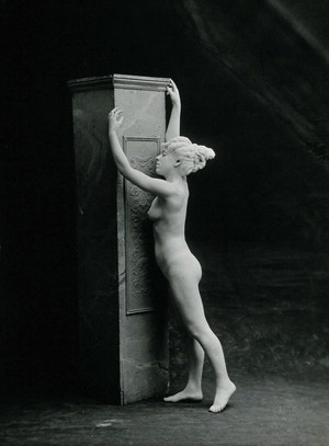 view A young woman, posing naked in a classical attitude, against a square column in a photographic studio. Photograph, ca.1890/1910.