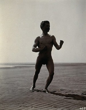 view A man posing naked in a boxing stance with his fists raised, on a deserted beach.